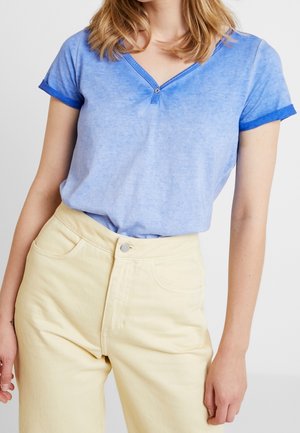 Femme portant un t-shirt bleu clair à col en V avec des poignets de manches bleu foncé et un pantalon taille haute jaune pâle, debout devant un fond uni.