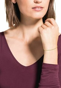 Pulsera de cadena de oro con eslabones pequeños llevada en una muñeca. El fondo presenta una blusa burdeos intenso y una textura de piel suave.