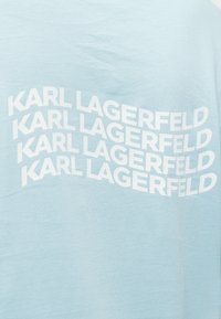 Ανοιχτό μπλε ύφασμα με την επιγραφή "KARL LAGERFELD" τέσσερις φορές σε λευκό, κάθε γραμμή ελαφρώς κυματιστή και στοιβαγμένη κάθετα.