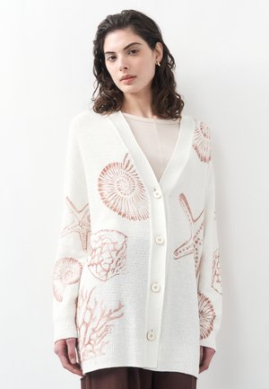 Woman with dark curly hair wearing a white buttoned cardigan featuring coral, seashell, and starfish designs.