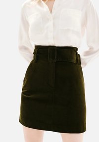 Femme portant une chemise blanche à boutons rentrée dans une jupe en velours côtelé vert foncé taille haute avec une large ceinture et une boucle rectangulaire.