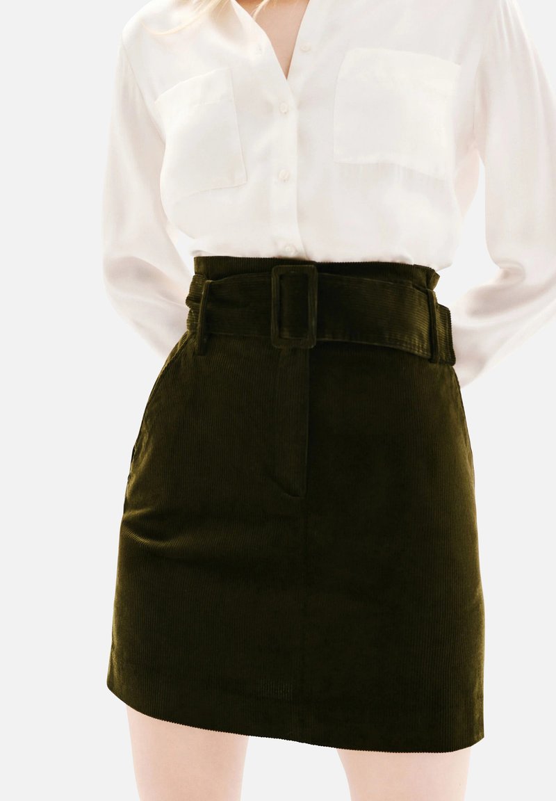 Femme portant une chemise blanche à boutons rentrée dans une jupe en velours côtelé vert foncé taille haute avec une large ceinture et une boucle rectangulaire.