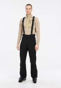 Pantalones de esquí negros con tirantes ajustables, combinados con una camiseta de manga larga beige con cuello con cremallera y acentos amarillos en contraste.