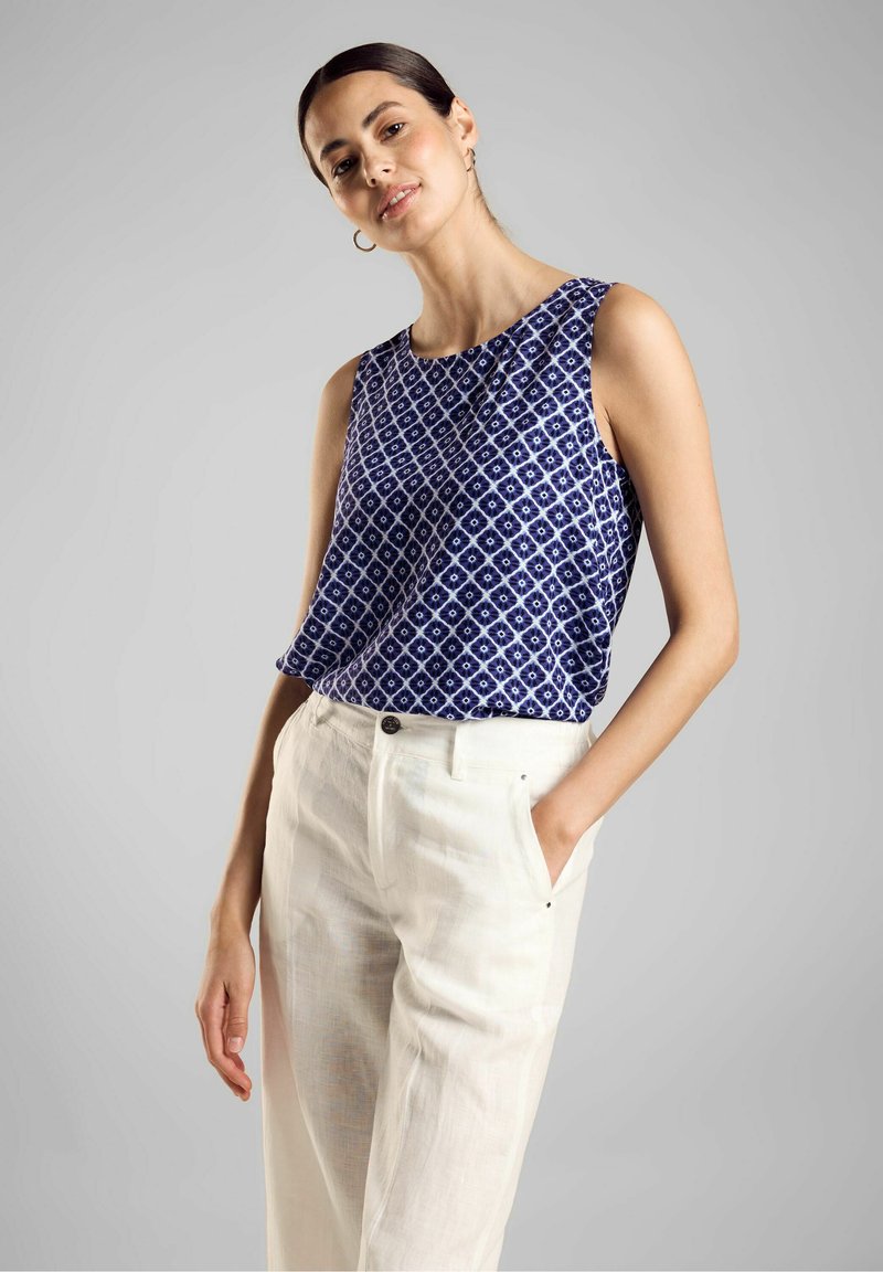 Top senza maniche blu navy con un motivo geometrico bianco, abbinato a pantaloni di lino chiari. Il tessuto sembra liscio con una vestibilità rilassata.