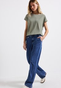 Světle zelené tričko s krátkým rukávem a nápisem "CONNECT", spárované s volnými modrými džíny. Model stojí, má ruce v kapsách a na sobě bílé tenisky.