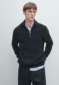 Dunkelblauer, gerippter Strickpullover mit einem hochschließenden Reißverschlusskragen, getragen über einem hellblauen Hemd mit Kragen, kombiniert mit dunklen Denim-Jeans.