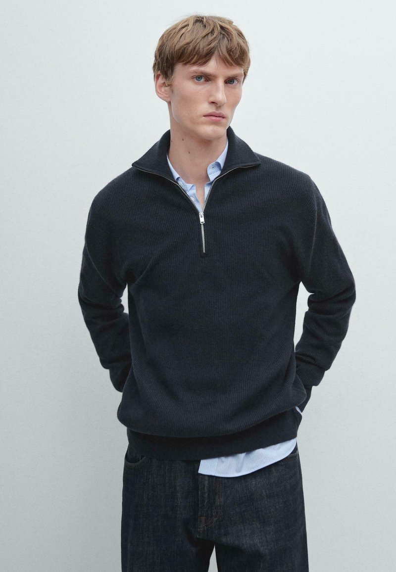 Dunkelblauer, gerippter Strickpullover mit einem hochschließenden Reißverschlusskragen, getragen über einem hellblauen Hemd mit Kragen, kombiniert mit dunklen Denim-Jeans.