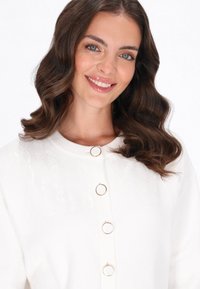 Weißer Cardigan mit einem strukturierten Obermaterial und einem Zopfmuster, runden goldfarbenen Knöpfen vorne und einem weichen, glatten Stoff.