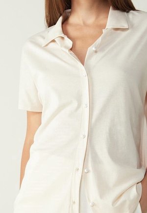 Femme portant une chemise crème à manches courtes, boutonnée avec col, mains partiellement dans les poches, sur un fond uni.