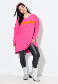Tunika lunga rosa a maniche lunghe con "GOOD VIBES" in arancione, abbinata a leggings neri lucidi e stivaletti d'angolo argentati. Tessuto liscio, vestibilità comoda.