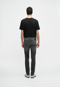 Άνδρας που στέ κεται με πλάτη στην κάμερα, φορώντας μαύρη μπλούζα, ξεθωριασμένα μαύρα τζιν με ορατές τσέπες και λευκά αθλητικά παπούτσια, σε απλό φόντο.