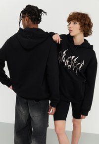 Zwarte hoodies van zacht materiaal; één heeft een metalen vlamontwerp op de voorkant. Beiden zijn gecombineerd met donkere jeans en shorts.