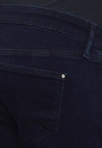 Jeans de ganga azul escuro com um design de cinco bolsos. Apresenta um pequeno logotipo perto do bolso traseiro e costuras em contrastante. Textura suave.