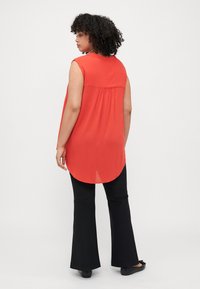 Haut rouge sans manches avec un ourlet arrondi, en tissu texturé, et détail froncé au dos, associé à un pantalon noir évasé et des chaussures plates.