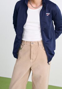 Laivaston sininen vetoketjullinen takki, jossa on Reebokin logo, yhdistettynä beigeihin korkeavyötäröisiin housuihin. Alla valkoinen ribattu toppi ja hopeinen ketjukaulakoru.