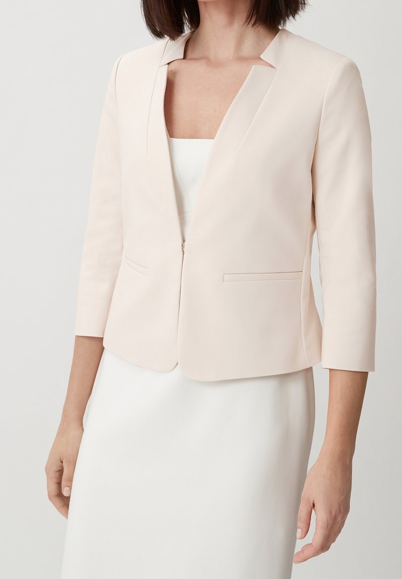 Femme portant un blazer court beige clair à manches trois-quarts sur une robe blanche, vue du cou jusqu'à mi-cuisse.