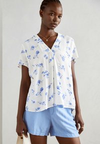 Blusa bianca a maniche corte con un motivo floreale blu, design a V, fit morbido. Abbinata a pantaloni corti azzurri, tiene una borsa color crema.
