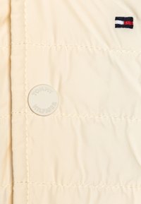 Beige quiltet jakke med en rund Tommy Hilfiger-logo og et lille broderet flagmærke i rød, hvid og blå.