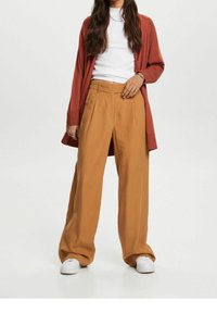 Cardigan couleur rouille porté sur une chemise blanche, associé à un pantalon large marron clair et des baskets blanches. Tissu lisse avec une coupe ample.