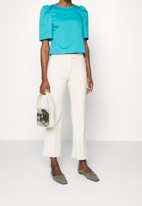 Blouse courte bleu sarcelle à manches bouffantes, associée à un pantalon crème ajusté. Le mannequin tient un sac à main transparent. Des ballerines grises complètent le look.