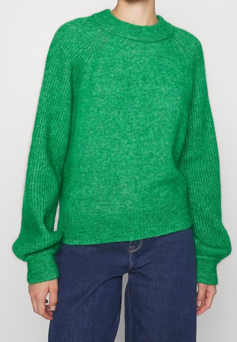 Grön stickad tröja med ribbad textur, rundad halsringning och voluminösa ärmar, tillsammans med mörka denimjeans.