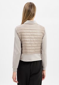 Lichtgewicht, beige puffer vest met horizontale stiksels, een kraaghalslijn en contrasterende mouwen, gecombineerd met zwarte broek.