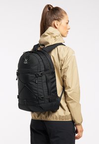 Mochila negra con respaldo acolchado y correas ajustables, con múltiples compartimentos con cremallera y acentos de cordón elástico. Llevada con una chaqueta beige.