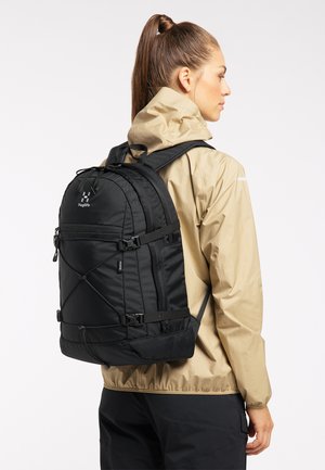 Schwarzer Rucksack mit gepolstertem Rücken und verstellbaren Riemen, ausgestattet mit mehreren Reißverschlussfächern und Akzenten aus Schnur. Getragen mit einer beigen Jacke.