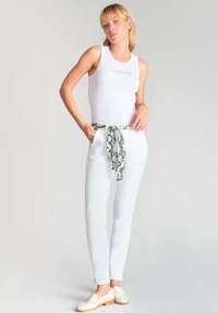 Weißes geripptes Tanktop, weiße taillierte Hose mit einem Schlangenprint-Schal als Gürtel und beigefarbene Loafers, vor einem neutralen Hintergrund.