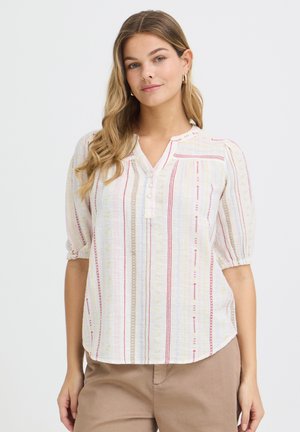 Kvinde iført en lys bluse med lodrette røde og beige striber og beige bukser, stående mod en ensfarvet hvid baggrund.