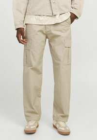 Beige cargo bukser lavet af bomuld, med flere lommer, en afslappet pasform og synlige syninger. Båret med beige sneakers.