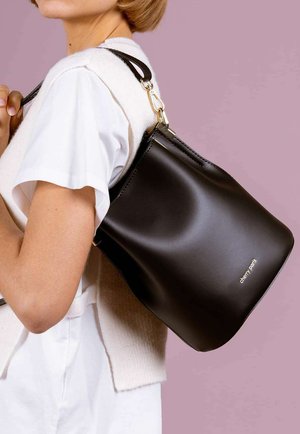 Femme portant un t-shirt blanc et un gilet, portant un sac seau en cuir noir avec une fermeture dorée et le logo "cherry paris" sur l'épaule, sur un fond rose.