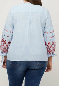 Blouse rayée bleue avec des accents floraux rouges brodés sur les manches bouffantes, présentant des liens aux poignets et un dos uni.