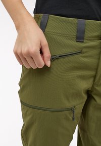 Olivově zelené kalhoty s kapsou na zip a texturovanou tkaninou. Pás obsahuje šedé akcenty a detaily šití pro větší odolnost.