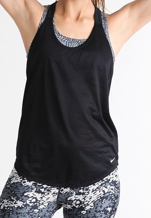 Mujer con camiseta sin mangas negra y leggings con patrón en blanco y negro, brazos levantados contra un fondo liso.