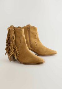 Botines de ante en color arena con punta afilada, cremallera lateral y detalles de flecos en la parte trasera, que cuentan con un tacón de madera apilado.