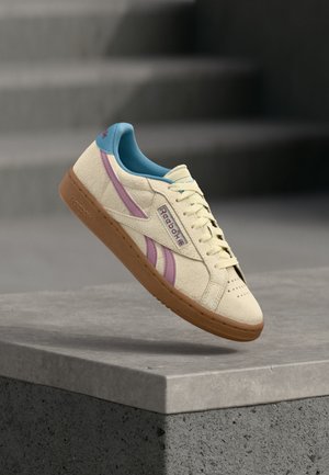 Kremasto Reebok tenisica s ružičastim prugama, plavim unutarnjim postavom i smeđim potplatom lebdi iznad betonske ograde.