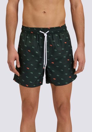 Uomo che indossa pantaloncini da bagno neri con piccoli motivi di foglie verdi e arancioni e un cordoncino bianco legato in vita.