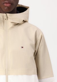 Beige hooded jacket with a smooth texture, featuring a white lower section and a small, colored logo. Zippered front closure.