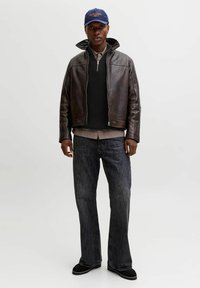 Braune Lederjacke mit hohem Kragen, schwarzer Strickpullover, kariertes Hemd, dunkelblaue Denim-Jeans und schwarze Schuhe. Blaue Kappe mit gesticktem Detail.