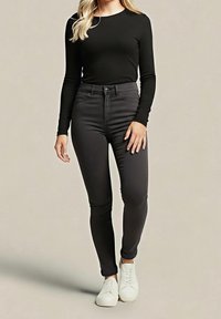 Femme portant un haut noir à manches longues, un jean skinny taille haute foncé et des baskets blanches, debout devant un fond uni.
