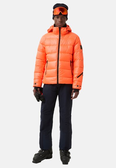 Jasnopomarańczowa puchowa kurtka z zamkiem z przodu, czarnymi akcentami i kapturem, zestawiona z ciemnoniebieskimi spodniami narciarskimi i czarnymi butami narciarskimi.