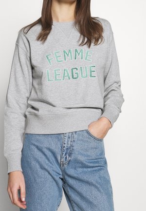 Kobieta ubrana w szarą bluzę z zielonym tekstem "FEMME LEAGUE" i niebieskie jeansy z wysokim stanem, stoi z jedną ręką w kieszeni.