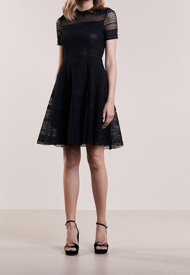 Femme portant une robe en dentelle noire longueur genou avec des manches courtes et des sandales noires à talons hauts avec brides à la cheville.