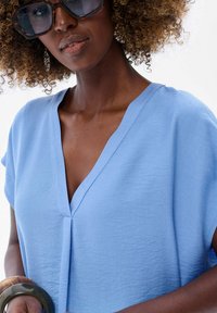 Jasnoniebieska bluzka z krótkimi rękawami i dekoltem w kształcie litery V, wykonana z teksturowanej tkaniny. Zawiera środkowy fałd i minimalistyczny styl.