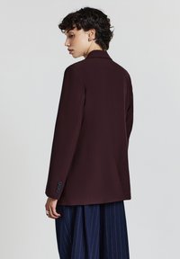 Blazer bordeaux con un design strutturato, maniche lunghe, chiusura a un bottone e tasche laterali, abbinato a pantaloni a righe.