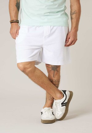 Homme portant un short de sport blanc, des baskets blanches avec des détails noirs, un t-shirt bleu clair, debout avec les jambes croisées, tatouages visibles sur les bras et les jambes.