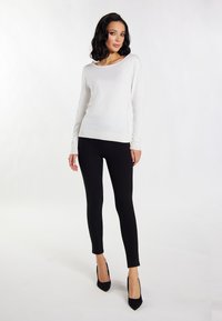 Top alb cu mâneci lungi și accente texturate, asortat cu leggings negri obținuți pe corp și tocuri stiletto negre, pe un fundal neutru.