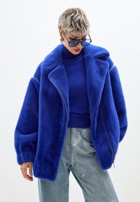 Giacca in pelliccia sintetica blu con vestibilità rilassata, colletto oversize e chiusura con zip, abbinata a un top blu e pantaloni di denim chiaro.