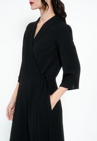 Fekete, háromnegyedes ujjú bodycon ruha, oldalsó megkötővel és diszkrét zsebbel. Sima textúrájú és elegáns dizájnú.